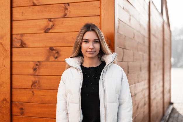 Young blonde woman in a black sweater and a fashionable white jacket posing near wooden building