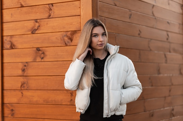 Young blonde woman in a black sweater and a fashionable white jacket posing near wooden building