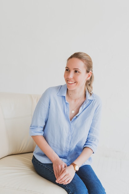 Young blonde woman in a blue shirt sits on a sofa in a bright room and laughs