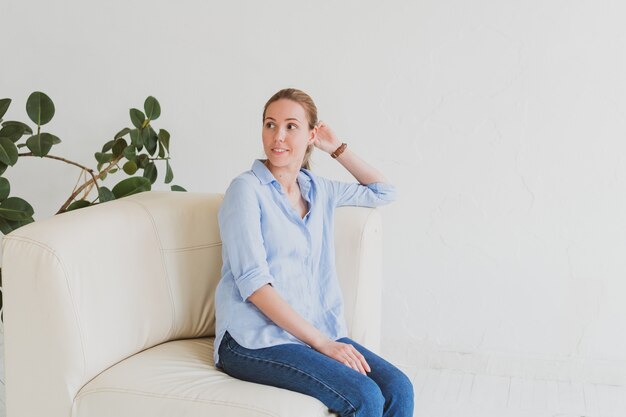 Young blonde woman in a blue shirt sits on a sofa in a bright room and smiles.