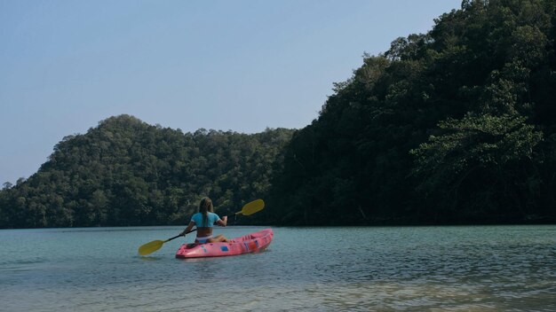 Young blonde woman in blue swimsuit rows pink plastic canoe along azure sea bay past island with palms under blue sky at resort. Traveling to tropical countries. Girl is sailing on kayak in ocean.