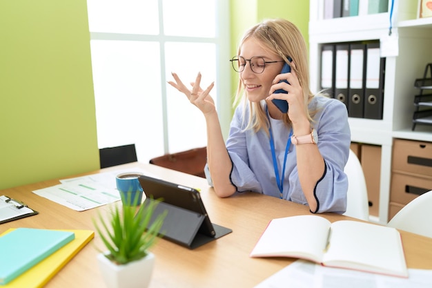 Young blonde woman business worker using touchpad talking on smartphone at office