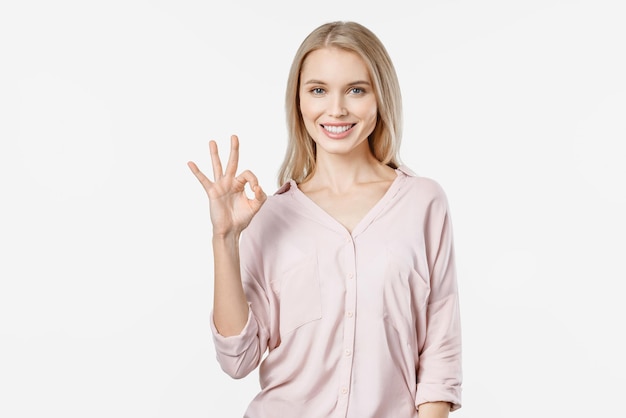 Young blonde woman in casual showing OK gesture and looking at camera isolated on white background