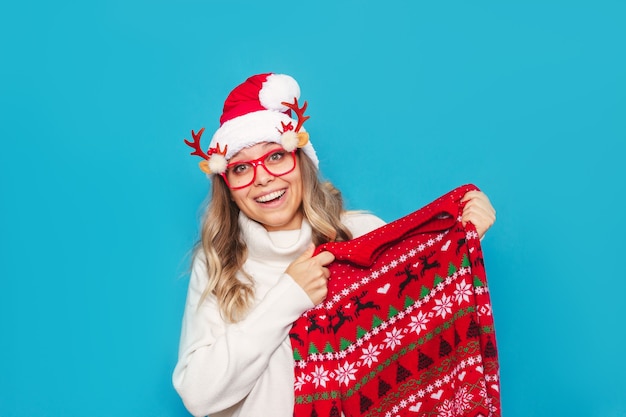 A young blonde woman in Christmas Santa hat and sweater holds red sweater with deer pattern