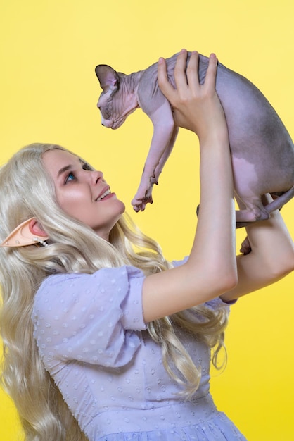 Young blonde woman cosplay elf in blue dress holds sphinx kitten in hands high above her head