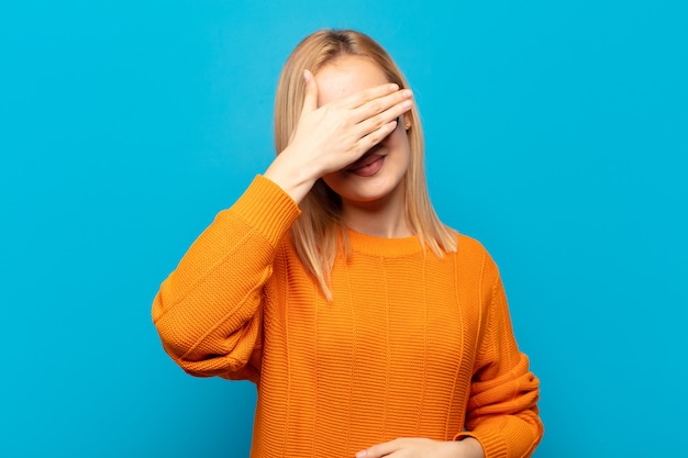 Young blonde woman covering eyes with one hand feeling scared or anxious, wondering or blindly waiting for a surprise