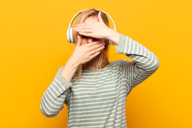 Giovane donna bionda che copre il viso con entrambe le mani dicendo no alla telecamera! rifiutare le foto o vietare le foto