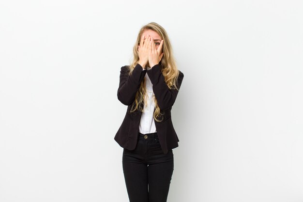 Young blonde woman covering face with hands, peeking between fingers with surprised expression and looking to the side against white wall