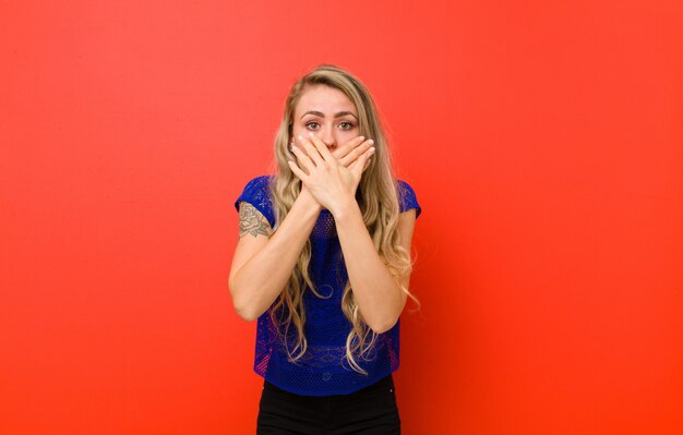 Young blonde woman covering mouth with hands with a shocked, surprised expression, keeping a secret or saying oops against red wall