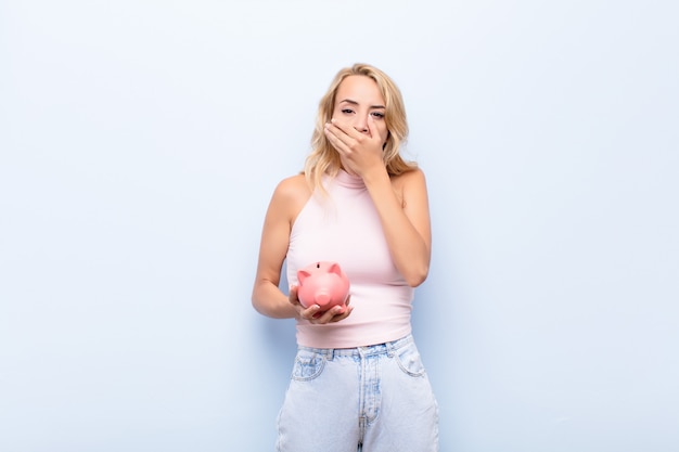 Young blonde woman covering mouth with hands with a shocked, surprised expression, keeping a secret or saying oops holding a piggybank