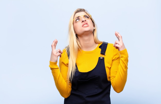 Young blonde woman crossing fingers anxiously and hoping for good luck with a worried look on flat wall