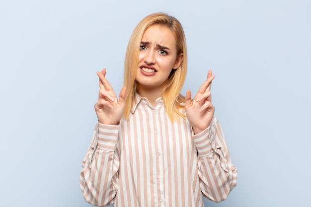 Young blonde woman crossing fingers anxiously and hoping for good luck with a worried look