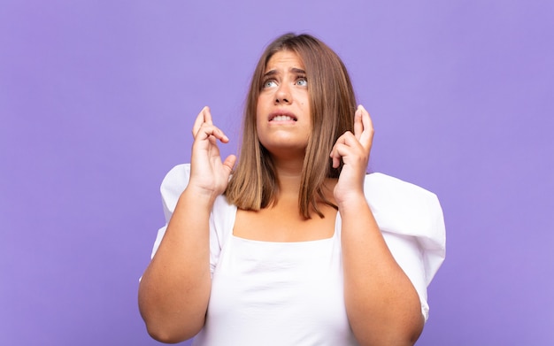 Young blonde woman crossing fingers anxiously and hoping for good luck with a worried look