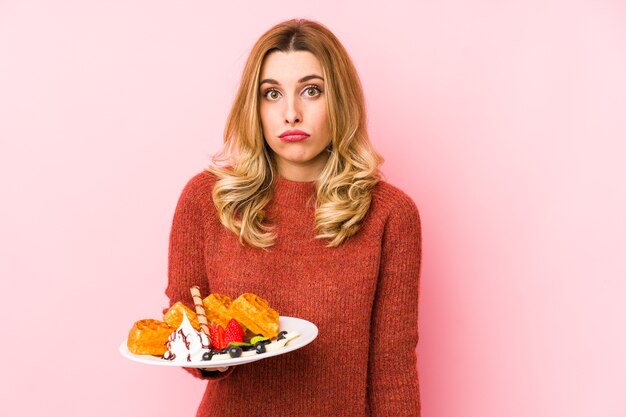 Young blonde woman eating a waffle dessert isolated shrugs shoulders and open eyes confused.