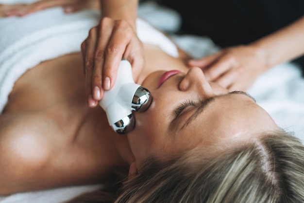 Young blonde woman enjoys facial massage with microcurrent facial massager in the spa