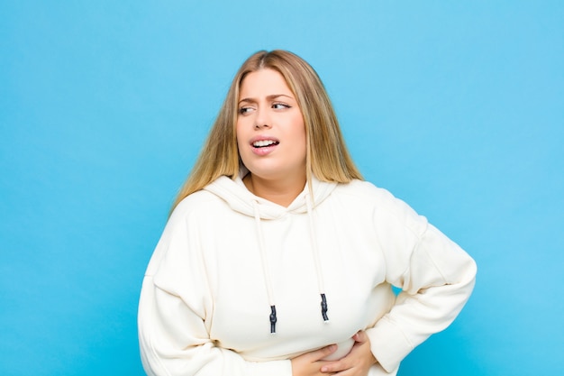 Young blonde woman feeling anxious, ill, sick and unhappy, suffering a painful stomach ache or flu against flat wall