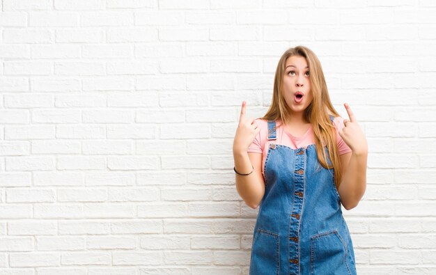 Young blonde woman feeling awed and open mouthed pointing upwards with a shocked and surprised look over brick wall