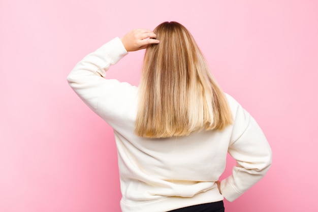 Young blonde woman feeling clueless and confused, thinking a solution, with hand on hip and other on head, rear view against flat wall