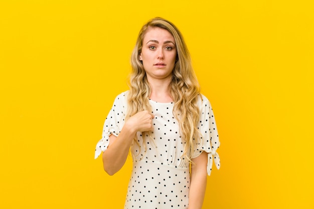 Young blonde woman feeling confused, puzzled and insecure, pointing to self wondering and asking who, me? against yellow wall