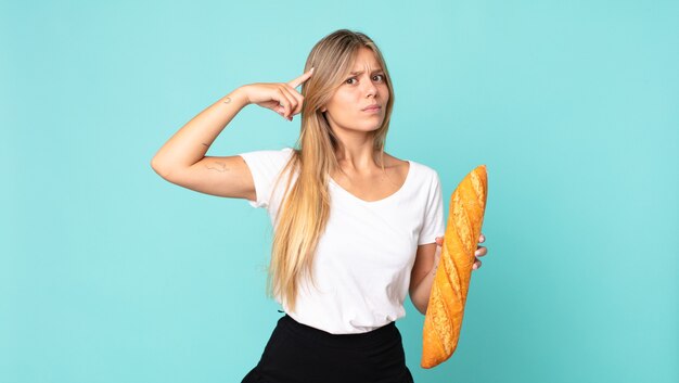 Young blonde woman feeling confused and puzzled, showing you are insane and holding a bread baguette