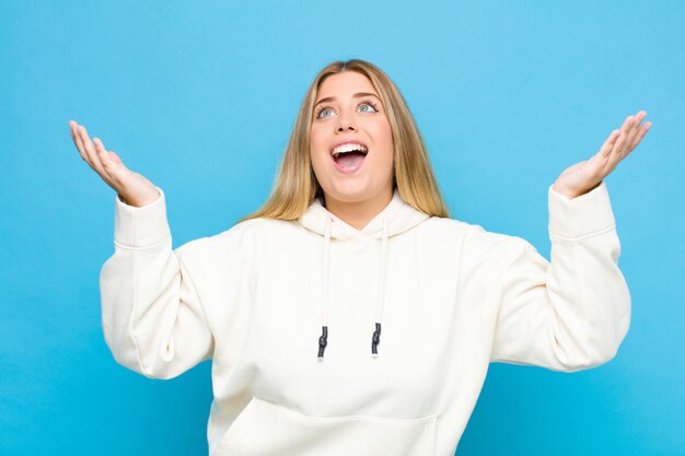 Young blonde woman feeling happy, amazed, lucky and surprised, celebrating victory with both hands up in the air over wall