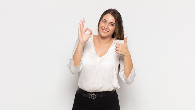 Giovane donna bionda che si sente felice, stupita, soddisfatta e sorpresa, mostrando i gesti ok e il pollice in alto, sorridente