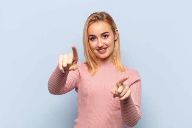 Young blonde woman feeling happy and confident, pointing to front with both hands and laughing, choosing you