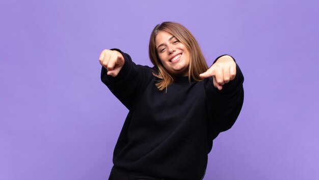 Young blonde woman feeling happy and confident, pointing with both hands and laughing, choosing you