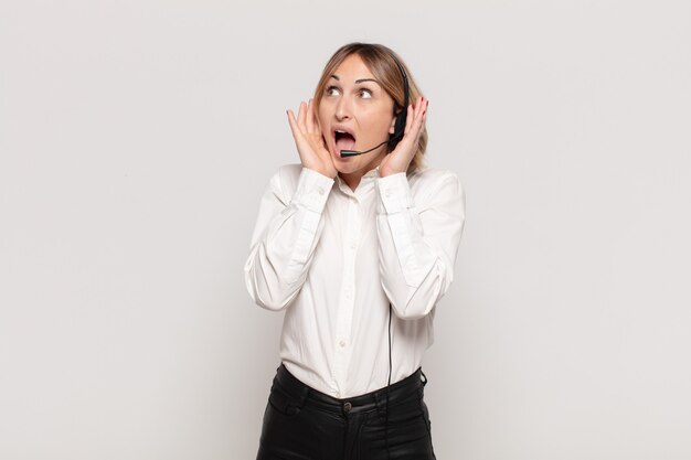 Young blonde woman feeling happy, excited and surprised, looking to the side with both hands on face