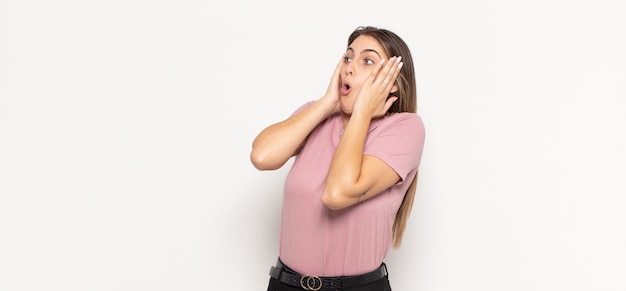 Young blonde woman feeling happy, excited and surprised, looking to the side with both hands on face