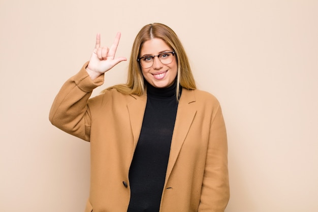 Young blonde woman feeling happy, fun, confident, positive and rebellious, making rock or heavy metal sign with hand against flat wall