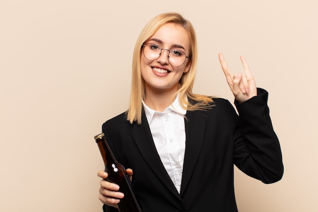 Young blonde woman feeling happy, fun, confident, positive and rebellious, making rock or heavy metal sign with hand