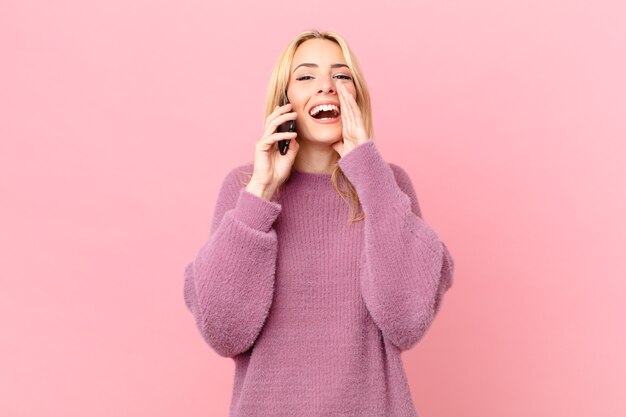 Young blonde woman feeling happy,giving a big shout out with hands next to mouth and talking with a smartphone