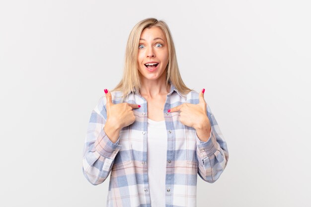 Young blonde woman feeling happy and pointing to self with an excited