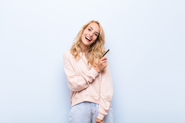 Young blonde woman feeling happy, positive and successful, motivated when facing a challenge or celebrating good results holding a credit card