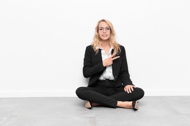 Young blonde woman feeling happy, positive and successful, with hand making v shape over chest, showing victory or peace sitting on the floor
