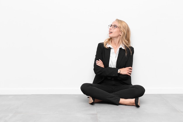 Photo young blonde woman feeling happy, proud and hopeful, wondering or thinking, looking up to copy space with crossed arms sitting on the floor