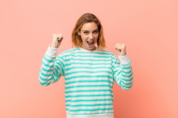 Young blonde woman feeling happy, surprised and proud, shouting and celebrating success with a big smile against flat color wall