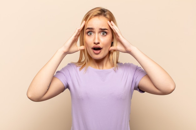 Young blonde woman feeling horrified and shocked, raising hands to head and panicking at a mistake