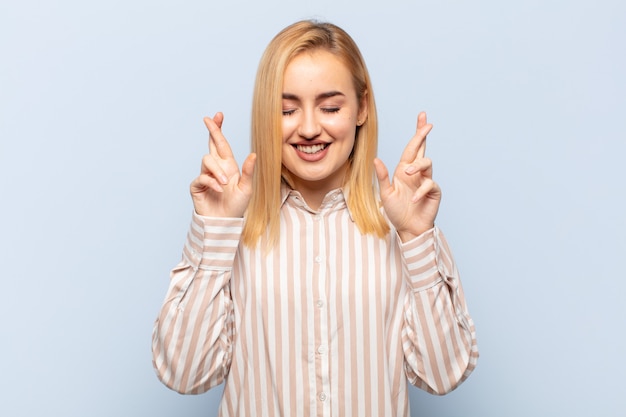 Young blonde woman feeling nervous and hopeful, crossing fingers, praying and hoping for good luck