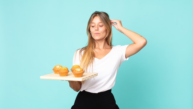 Young blonde woman feeling puzzled and confused, scratching head and holding a muffins troy