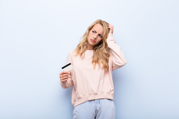 Young blonde woman feeling puzzled and confused, scratching head and looking to the side holding a credit card