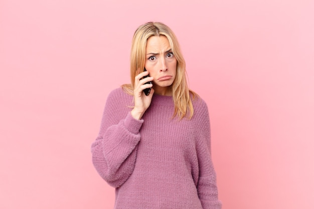 Young blonde woman feeling sad and whiney with an unhappy look and crying and talking with a smartphone