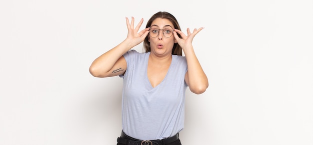 Young blonde woman feeling shocked, amazed and surprised, holding glasses with astonished, disbelieving look