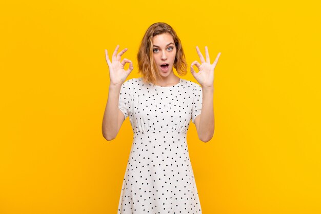 Young blonde woman feeling shocked, amazed and surprised, showing approval making okay sign with both hands against flat color wall