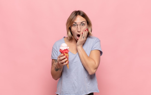 Young blonde woman feeling shocked and scared, looking terrified with open mouth and hands on cheeks