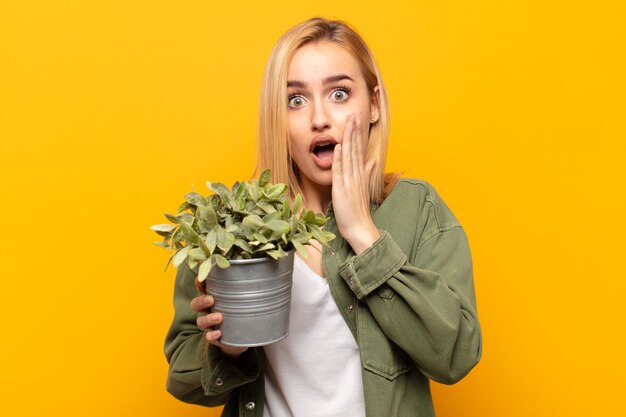 Young blonde woman feeling shocked and scared, looking terrified with open mouth and hands on cheeks