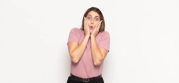 Young blonde woman feeling shocked and scared, looking terrified with open mouth and hands on cheeks