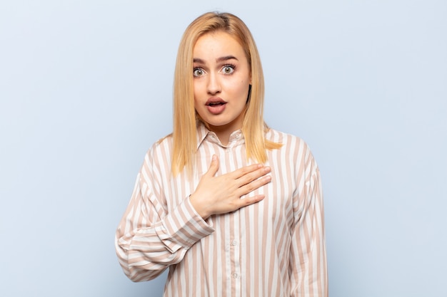 Young blonde woman feeling shocked and surprised, smiling, taking hand to heart, happy to be the one or showing gratitude