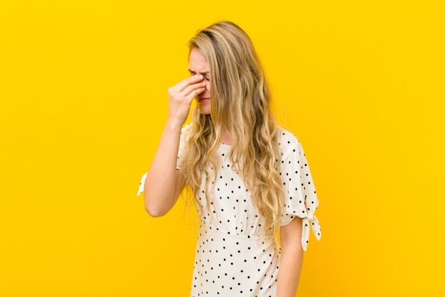 Young blonde woman feeling stressed, unhappy and frustrated, touching forehead and suffering migraine of severe headache against yellow wall